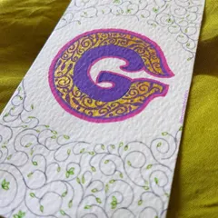 An illuminated letter 'G' decorated with intricate gold details and delicate vine-like flourishes. This style of work is perfect for creating custom monograms for wedding invitations or personalized stationery.