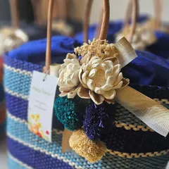 Another variation of the raffia basket, this one featuring a striped blue pattern. The consistent floral and pom-pom detailing ensures a cohesive look for large orders while allowing for slight variations in color.