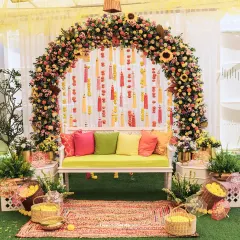 This Mehendi seating is a burst of color, featuring a floral arch with hanging tassels, a comfortable bench with bright cushions, and lush greenery, creating a perfect photo-ready spot.