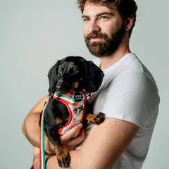 Capturing the bond between a pet and their person can make a product feel more relatable. Here, a model holds a sweet dachshund wearing the "Ciao Bella" harness from Luxolandshop, creating a warm, lifestyle-oriented studio portrait.