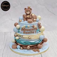A sweet two-tier "Adventure Awaits" cake to welcome a new baby. This 3kg choco-chip cake features a crowned teddy bear topper amidst blue, brown, and gold decorative balls and macarons.