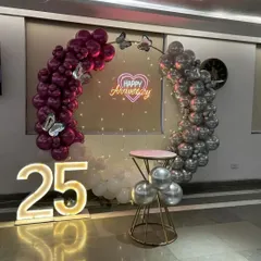 Celebrating a silver jubilee with a stylish balloon arch in magenta and silver. This 25th-anniversary setup includes large light-up numbers and a "Happy Anniversary" neon sign, perfect for photos.
