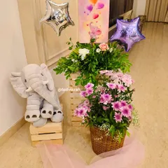 A sweet corner setup for a baby girl's arrival. It features a plush elephant toy, star-shaped balloons, and baskets of fresh pink and white flowers, creating a soft and celebratory welcome.