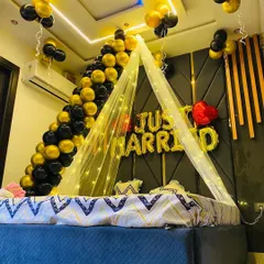 A chic and modern "Just Married" bedroom decoration featuring a black and gold balloon garland, a delicate fairy light canopy over the bed, and matching balloons floating on the ceiling. It's a stylish way to welcome the newlyweds home.
