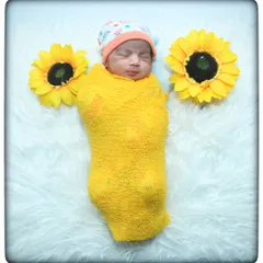 A little ray of sunshine. This newborn is wrapped in a cheerful yellow swaddle, sleeping peacefully between two large sunflower props on a soft white fur rug.