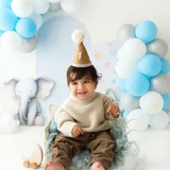 A smiling baby boy in a cozy sweater and party hat, celebrating his first birthday in the studio.