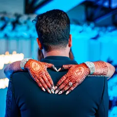 A powerful and emotional shot of the bride's henna-adorned hands making a heart shape. The back-hand design features the couple's initials and the engagement ring symbol, beautifully captured.