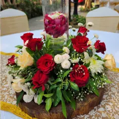 A classic centerpiece design with red and cream roses arranged on a rustic wooden slice. A tall glass vase in the center holds floating rose petals, adding a touch of modern elegance to the rustic base.