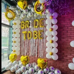 A fun 'Bride to Be' decoration against a brick wall. This setup uses gold foil letters, a white fringe curtain, and columns of daisy-shaped balloons made from white and gold balloons.