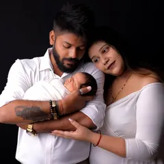 A complete family portrait with their newborn. The parents gaze down at their baby, wrapped snugly in white, creating a timeless image of love and unity against a classic black studio backdrop.