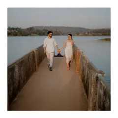 A couple walks hand-in-hand down a concrete pier over a lake, their coordinated white outfits creating a clean and classic look against the blue water.
