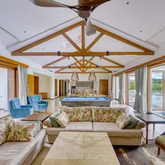The expansive living and entertainment area of the Kabini farmhouse. A high, vaulted ceiling with exposed wooden beams creates a sense of rustic grandeur, while large glass doors open up to the pool and landscape.