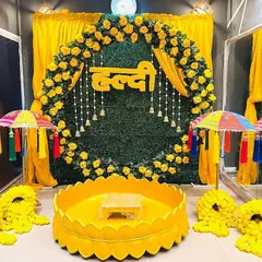 A classic Haldi ceremony backdrop featuring a lush green wall, a floral arch of yellow roses, and traditional bell hangings. The setup includes a central vessel for the rituals, all tied together with a vibrant yellow theme.