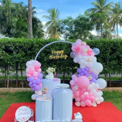 A bright and cheerful outdoor birthday decoration on a lawn, with a pastel balloon arch, white pedestals, and marquee letters.