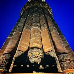 A dramatic low-angle shot of the Qutub Minar at night, emphasizing its fluted columns and detailed carvings against a deep blue sky.