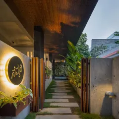 The entrance pathway is lined with stone pavers and lush greenery, leading you under a wood-clad portico. The illuminated nameplate, 'Nemmadi' (meaning peace or tranquility in Kannada), sets the tone for the entire home.