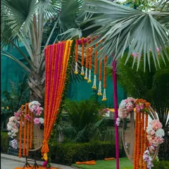 This is an outdoor entrance arch for a day event, set against lush greenery. The arch is decorated with strings of orange and pink marigolds, with floral arrangements on the side, creating a vibrant welcome.