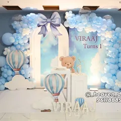 A dreamy hot air balloon and teddy bear theme for a first birthday, featuring a custom arched backdrop against a soft blue wall. The setup includes a full organic balloon arch in shades of blue and white, themed plinths, and large illuminated letters spelling out the name.