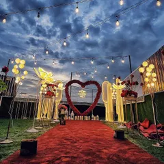 This wide-angle view shows a complete grand proposal aisle under a dramatic evening sky. The red carpet is flanked by unique illuminated floral sculptures and leads to a red rose heart arch, creating a spectacular entrance.