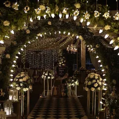 An elegant sangeet entranceway that combines a lush floral arch with warm bulb lighting and a classic checkerboard floor. This design creates a sophisticated and welcoming path for guests, perfect for a formal corporate event or gala.