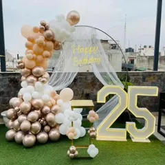 An elegant 25th birthday on a terrace with a beautiful arch of rose gold, beige, and white balloons. The setup includes a large '25' marquee light, perfect for an outdoor celebration.