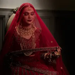 A candid moment under the veil. The soft lighting highlights the flawless base and the deep red tones of the makeup, creating a dreamy, romantic bridal portrait.
