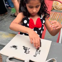 Little hands, big creativity. Here, a child is choosing and pressing wooden blocks onto her tote bag. Each stamp is a decision, a piece of a story she is creating, all while making a reusable bag to replace single-use plastics.