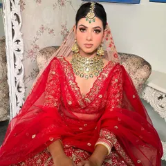 A regal portrait of a bride in a classic red lehenga. The makeup features sharp, defined eyes and a perfectly sculpted base, paired with a heavy kundan necklace to create a look of pure bridal opulence.