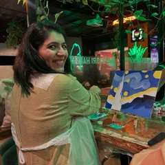 A participant smiling back at the camera, her version of 'Starry Night' in progress. The green neon lights of the cafe add to the cool, creative ambiance.