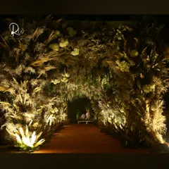 A magical walkway for a cocktail event, created with an arch of pampas grass and dried foliage, illuminated from below to create a warm, golden tunnel.