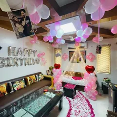 A full room decoration for a birthday surprise. The setup includes balloons on the ceiling, a "Happy Birthday" foil banner, and a path of rose petals leading to the decorated bed.