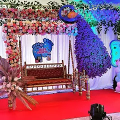 A vibrant peacock-themed decoration for a Dohale Jevan. The setup includes a floral swing, a large peacock figure made from purple flowers, and gender-themed cutouts with pink and blue balloons.
