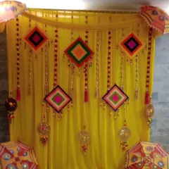 A festive Haldi backdrop with a bright yellow curtain and colorful hanging decorations. The design incorporates Rajasthani umbrellas and dreamcatchers for a vibrant, folk-art inspired theme.