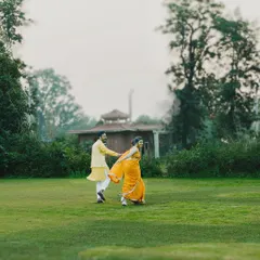 A couple shares a joyful run across a vast green lawn during their Haldi ceremony. The vibrant traditional outfits contrast beautifully with the natural setting, capturing the energy of the celebration.