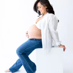A relaxed solo maternity pose in a casual outfit of jeans and a white shirt. This image shows a simple, modern approach to pregnancy portraits against a clean white studio background.