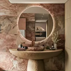 We crafted this statement bathroom to feel like a private spa. The design is centered around a custom pedestal sink and a backlit circular mirror, set against a dramatic wall of rose-toned natural stone, showcasing our expertise in luxury bathroom interiors and material selection.
