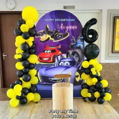 A Batman and Hot Wheels theme for Avyaan's 6th birthday. The decoration includes a custom printed backdrop with cars, black and yellow balloon columns, and a large number 6 foil balloon.