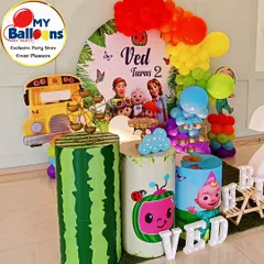 A complete Cocomelon-themed second birthday setup. This vibrant decor includes a rainbow balloon arch, the iconic yellow school bus, character-printed plinths, and a large light-up number '2'.