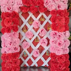 A compact backdrop design featuring a frame of red and pink artificial carnations. The background uses a white lattice pattern over red fabric, decorated with hanging golden bells and pom-poms.
