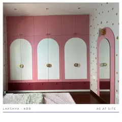 The centerpiece of this girl's room is the custom wardrobe, featuring a playful design with pink and white arched panels and unique gold hardware. The heart-patterned wallpaper adds a touch of whimsy.