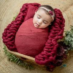 A peaceful portrait of a sleeping baby girl, swaddled in a rich maroon wrap and nestled in a chunky knit blanket.