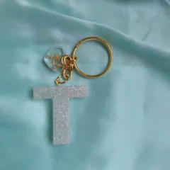 A simple and elegant initial keychain. This 'T' is made with a sparkling white glitter resin and comes with a small, clear heart charm filled with gold flakes, attached to a gold-plated ring.