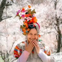 The joyful and shy smile of a Brokpa woman in Aryan Valley, Ladakh. Her vibrant floral headdress stands out against the soft white apricot blossoms, a perfect picture of spring.