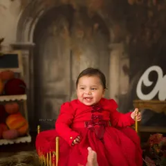 The birthday girl on her throne. Her bright red dress pops against the warm, rustic backdrop, and her smile is absolutely radiant as she celebrates turning one.