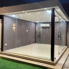 A modern, freestanding glass room set in a garden. This structure, with its minimalist frame and floor-to-ceiling glass walls, can serve as a home office, gym, or quiet retreat.