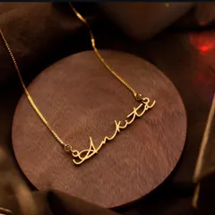 The 'Ankita' script necklace, presented on a wooden base, highlights the warm glow of the gold finish. Each nameplate is carefully crafted to be a personal treasure.