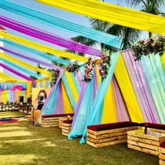 This outdoor Mehandi setup uses long, flowing drapes in pastel shades of yellow, blue, and purple to create a canopy. The low wooden seating with red cushions provides a comfortable and casual space for guests.