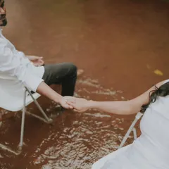 An artistic and symbolic shot of a couple holding hands while seated in chairs surrounded by water. This conceptual photo is about connection and trust.