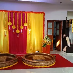 This is a classic Haldi ceremony setup for a home function. The backdrop is created with bright yellow and pink drapes, hanging marigold tassels, and traditional brass 'urlis' for the ceremony.