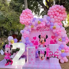 A perfect Minnie Mouse theme for a second birthday, set up outdoors. The decoration is all about pink, with a striped backdrop, large Minnie cutouts, and a beautiful balloon arch shaped like Minnie's ears.